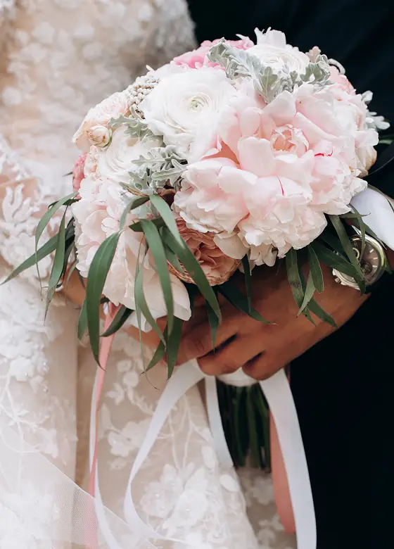 Bruidegom en bruid houden een pastelroze en witte bloemenboeket met groene bladeren vast. De kanten jurk van de bruid en het zwarte pak van de groom stralen elegantie uit.
