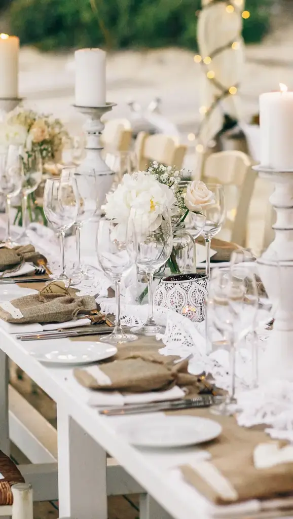 Elegante dinersetting aan het strand met een lange witte tafel versierd met kaarsen, beige linnen en bloemstukken. Warme, uitnodigende sfeer.