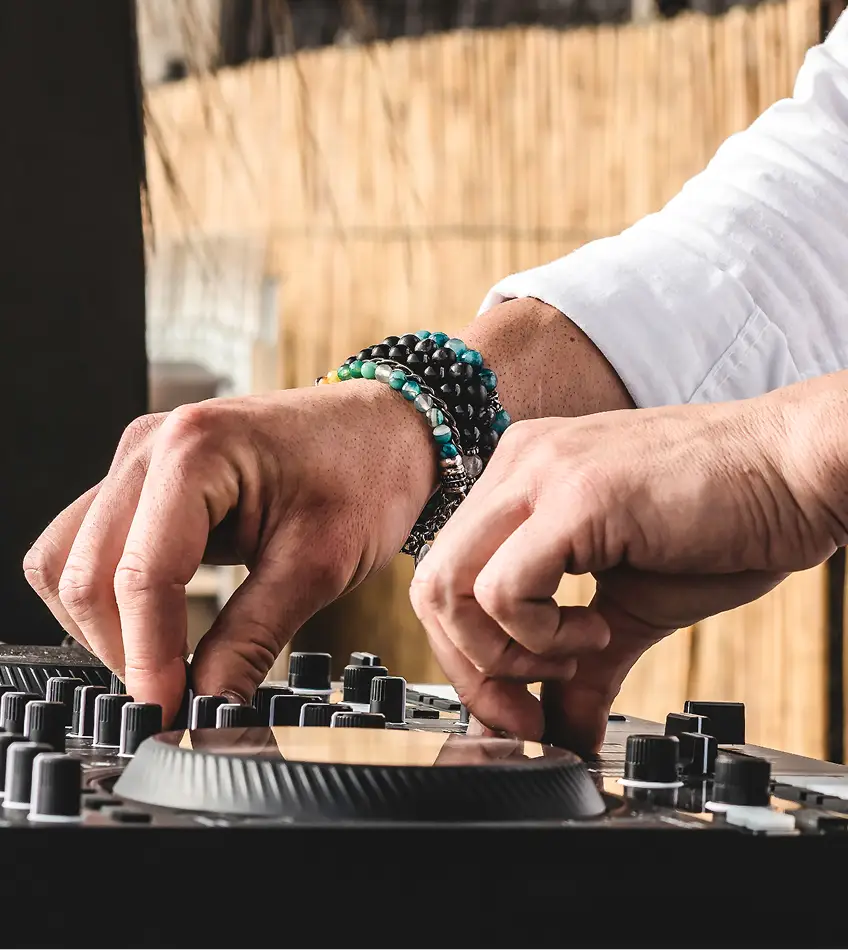 DJ in een wit shirt mixt vakkundig op een draaitafel, versierd met kleurrijke armbanden. Houten achtergrond, buitenomgeving. Energieke en gefocuste sfeer.