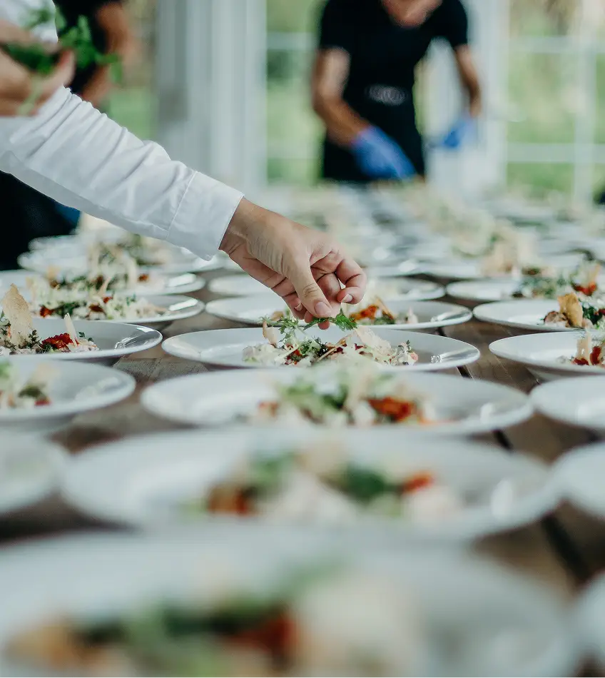 Chef-kok garneert rijen identieke borden met groenten in een heldere keuken, wat een gevoel van georganiseerde voorbereiding voor een groot evenement creëert.