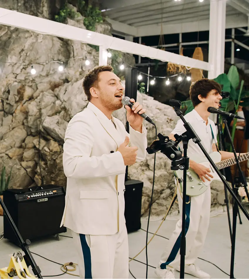 Drie muzikanten in witte trainingspakken met blauwe strepen performen energiek op het podium. Ze zingen in microfoons tegen een rotsachtige achtergrond met lichtsnoeren.