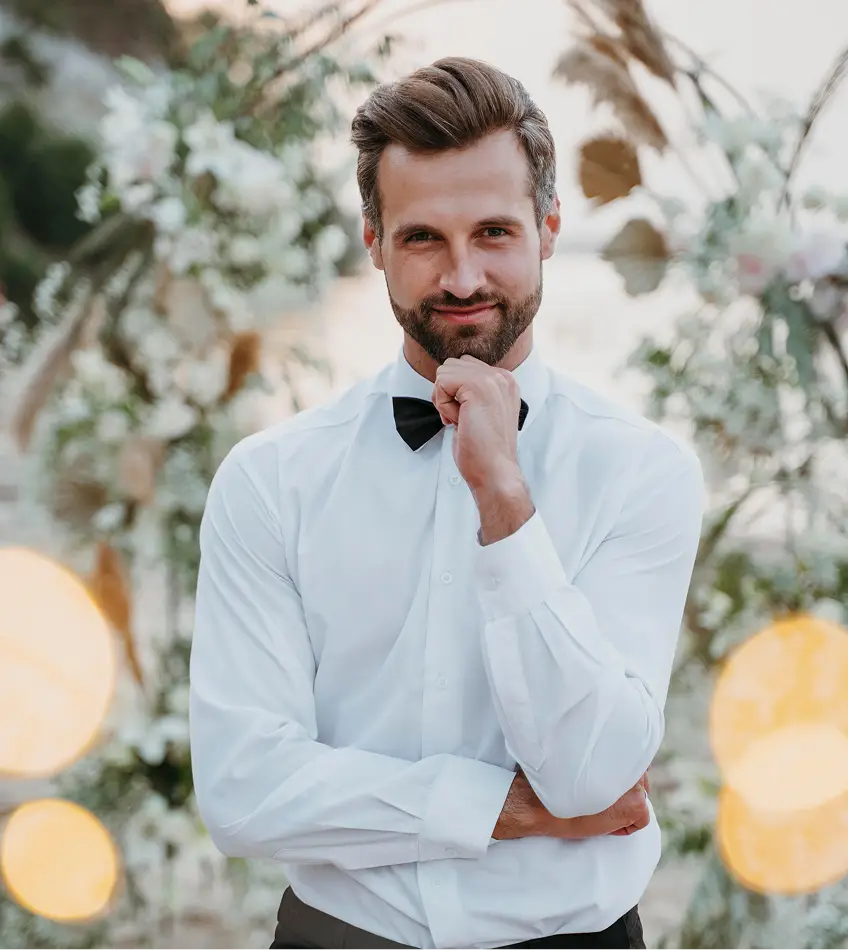 Een glimlachende man in een wit shirt en vlinderdas staat nadenkend op een strand, omringd door zachte bokehlichten en bloemarrangementen. Elegant en sereen.