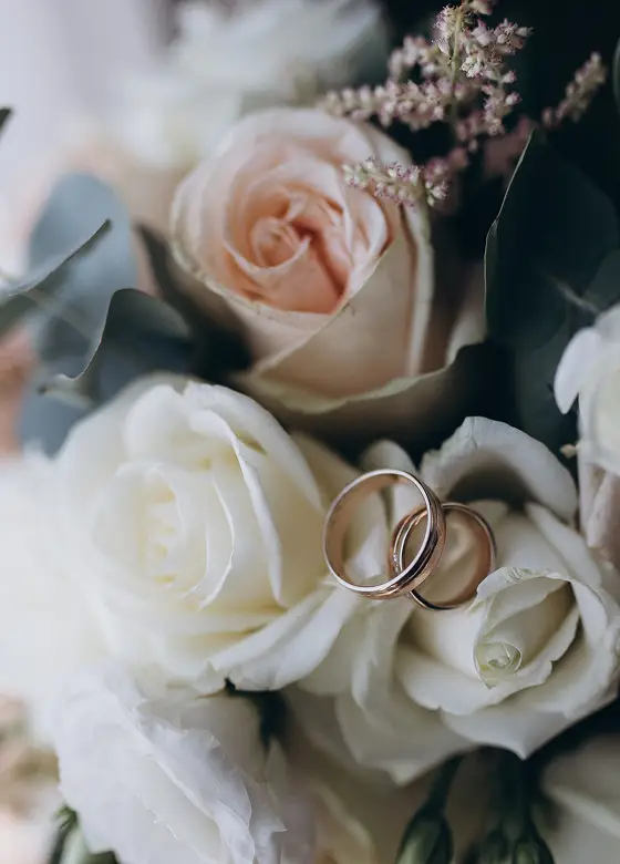 Een delicate boeket van witte en perzikroze rozen versierd met twee verstrengelde gouden trouwringen. Zachte, romantische sfeer met natuurlijk licht.